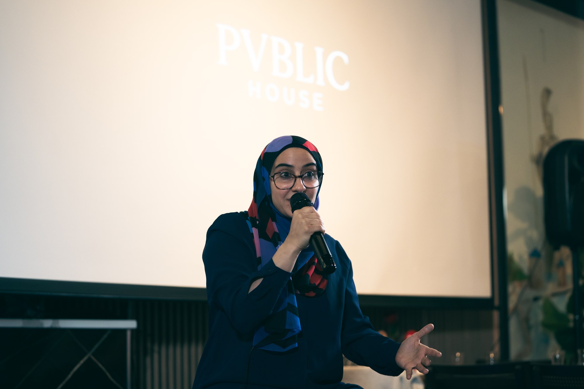 Speaker holding a microphone in front of a projected Pvblic House logo