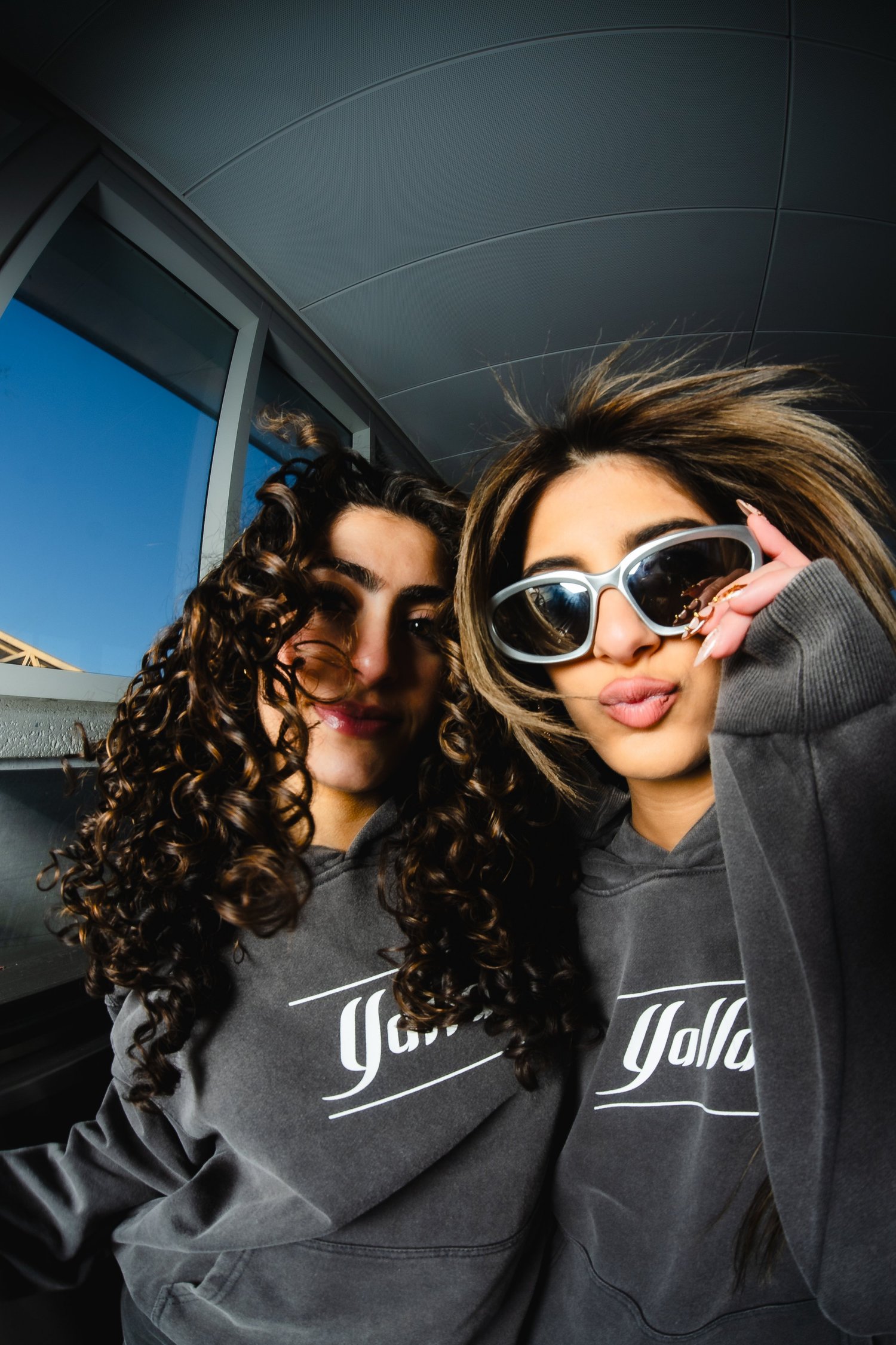 Two models wearing charcoal Yalla hoodies beside sunlit metro station windows